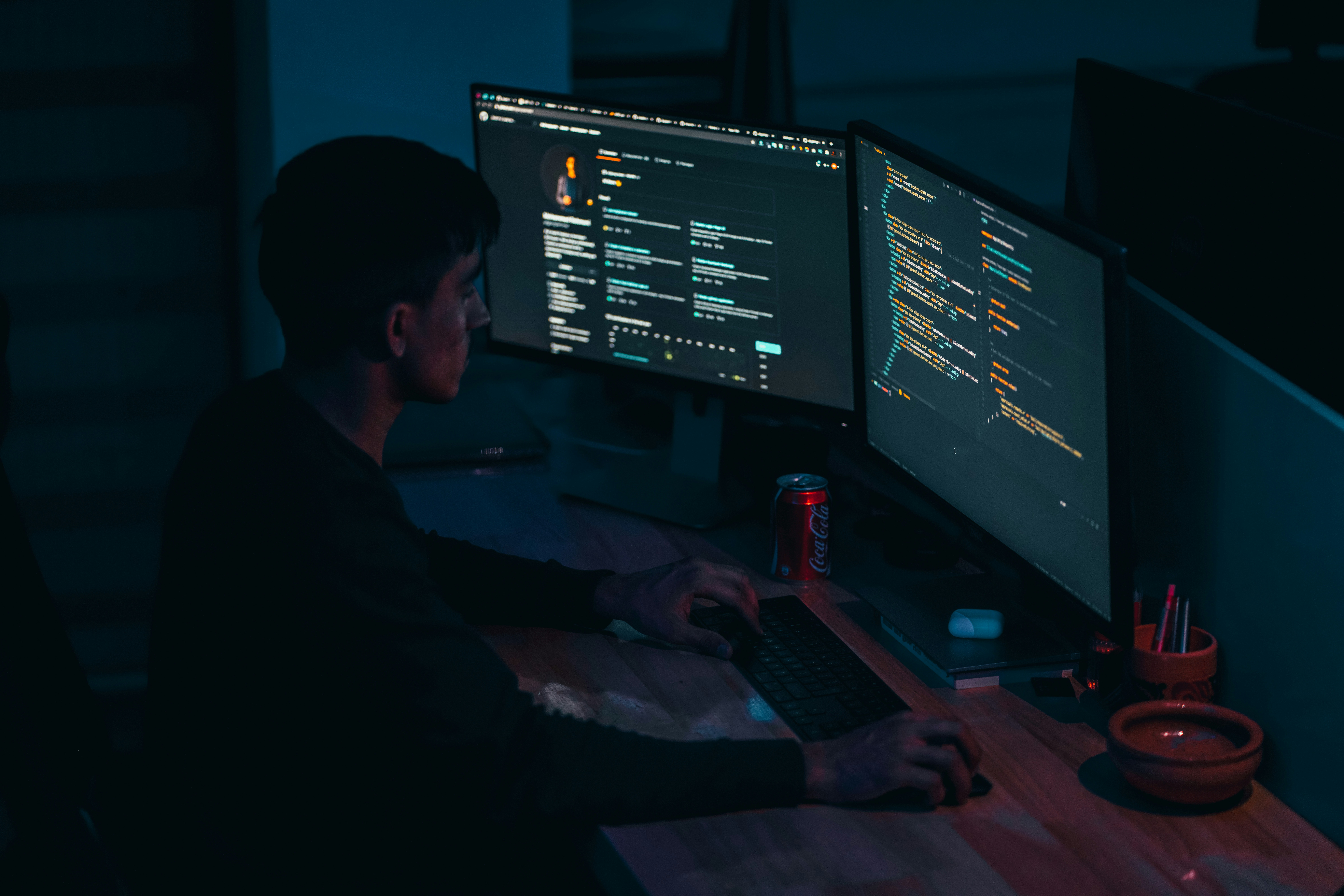 A developer working late at night at a dual-monitor setup, the room lit only by the blue glow of code on the screens